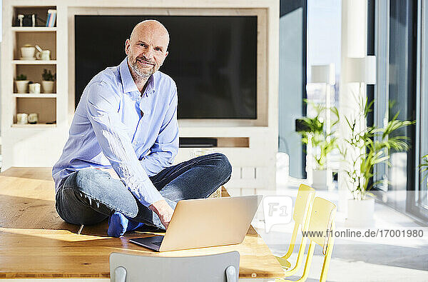 Lächelnder Geschäftsmann  der einen Laptop benutzt  während er zu Hause auf dem Tisch sitzt