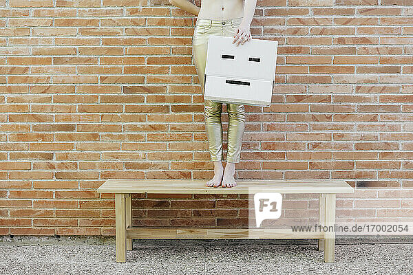 Frau steht auf einer Bank vor einer Backsteinmauer und hält eine Pappschachtel mit traurigem Gesicht