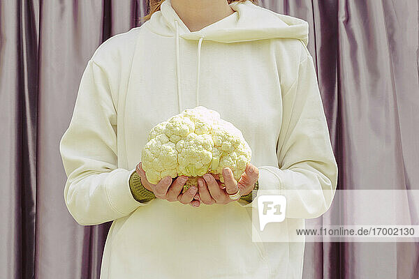 Close-up of mid adult woman holding cauliflower while standing against curtain at home