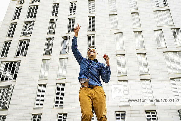 Fröhlicher Mann  der mit erhobener Hand gegen ein weißes Gebäude springt