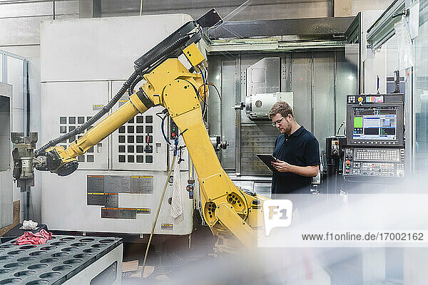 Männlicher Fabrikarbeiter verwendet ein digitales Tablet  während er vor Roboterarmen in der Industrie steht