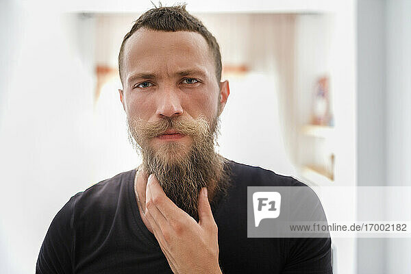 Männlicher Hipster berührt seinen Bart  während er zu Hause steht