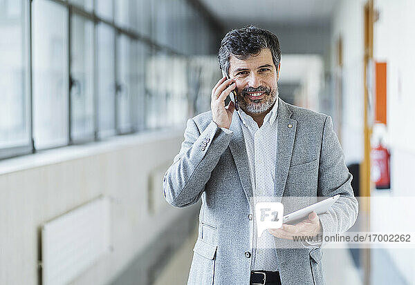 Glücklicher männlicher Professor  der ein Mobiltelefon benutzt und ein digitales Tablet im Flur einer Universität hält