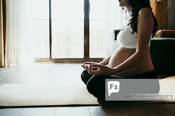 Schwangere Frau meditiert zu Hause auf einer Übungsmatte sitzend