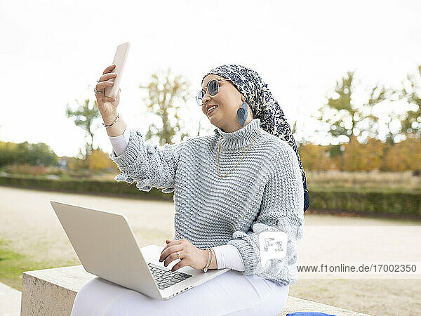 Lächelnde Frau mit Laptop  die ein Selfie macht  während sie im Freien sitzt
