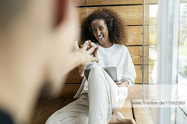 Playful woman with headphones and digital tablet stretching hand toward man while sitting at home