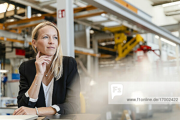 Lächelnde nachdenkliche Geschäftsfrau mit Hand am Kinn in einer Fabrik sitzend