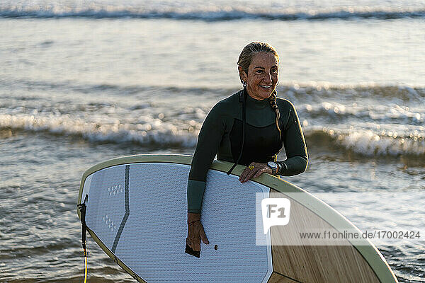 Smiling woman holding paddleboard standing in sea at dawn