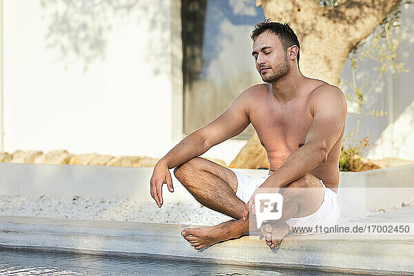 Hemdloser junger Mann in Shorts meditiert am Pool