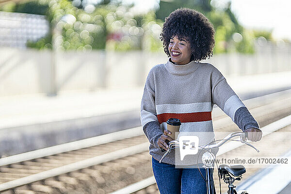 Nachdenkliche junge Frau mit wiederverwendbarem Becher auf dem Fahrrad am Bahnhof