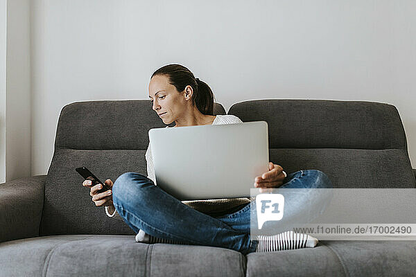Woman looking at smart phone while sitting with laptop on sofa