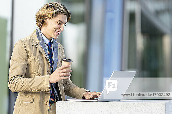 Smiling businessman with coffee cup working on laptop while standing outdoors