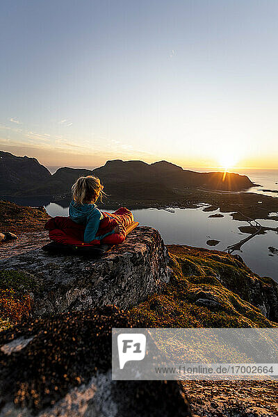 Frau bewundert die Aussicht auf den Sonnenuntergang  während sie im Schlafsack am Volandstinden  Lofoten  Norwegen  liegt