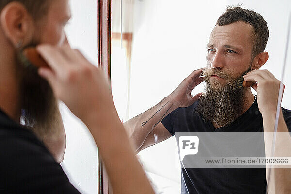 Mittlerer erwachsener Mann  der seinen Bart mit einem Kamm bürstet  während er einen Spiegel im Wohnzimmer betrachtet