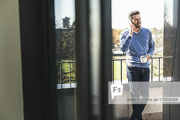 Geschäftsmann mit Kaffeetasse  der auf dem Balkon eines Büros steht und mit seinem Handy telefoniert
