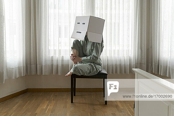Woman wearing a cardbox on head with bored smiley sitting on chair