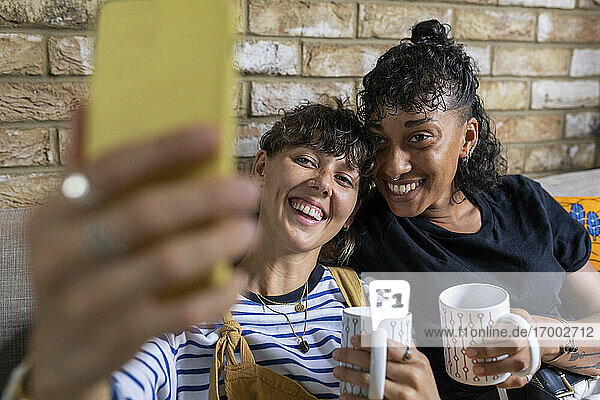 Junges lesbisches Paar mit Teetasse nimmt Selfie durch Handy  während zu Hause sitzen