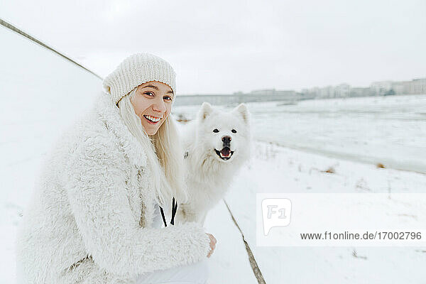 Portrait of white dressed smiling young woman with her white dog in the snow