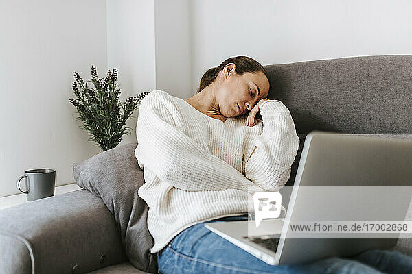Müde Frau schlafend mit Laptop auf Sofa im Wohnzimmer zu Hause sitzend
