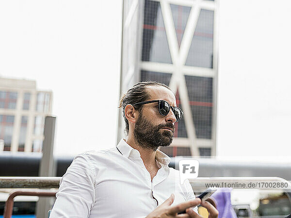 Portrait of bearded businessman wearing sunglasses sitting outdoors with smart phone in hands