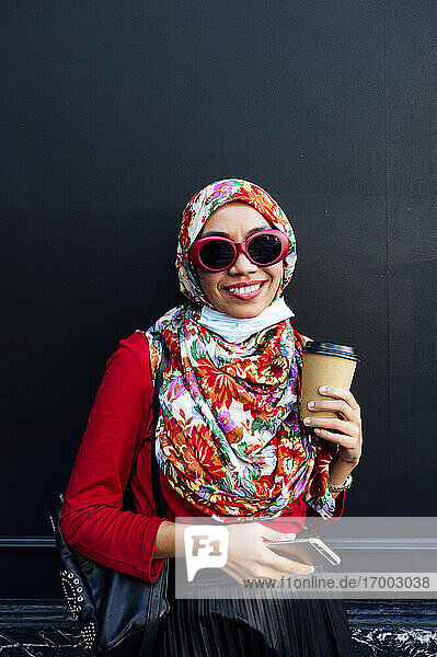 Lächelnde muslimische Frau hält Handy und Kaffeetasse gegen eine schwarze Wand während COVID-19