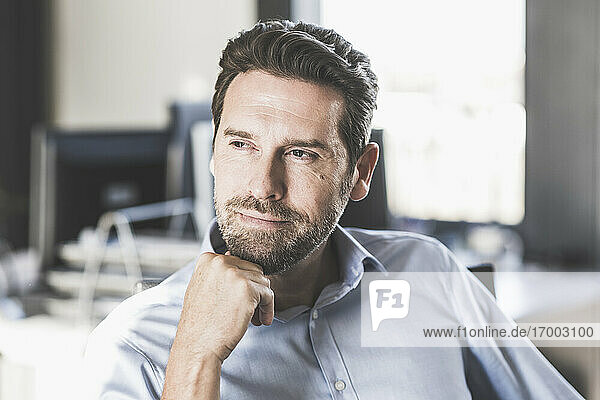 Thoughtful businessman with hand on chin looking away while sitting on chair at office
