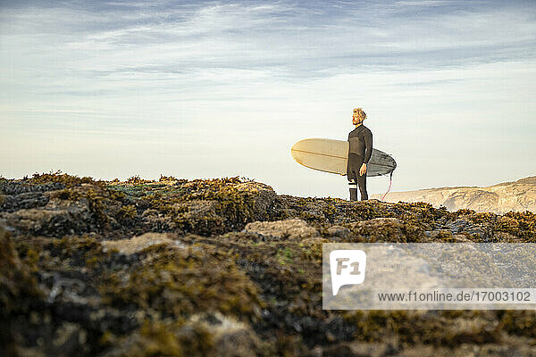 Male surfer standing with surfboard on rock formation at beach against sky