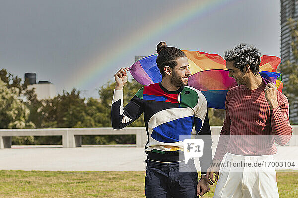 Lächelnder  liebevoller Mann mit Regenbogenschal  der seinen männlichen Partner in einem öffentlichen Park anschaut