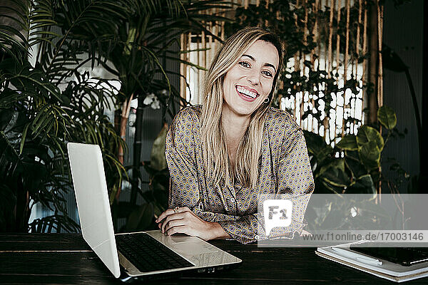 Glückliche weibliche Fachkraft sitzt mit Laptop am Schreibtisch im Büro