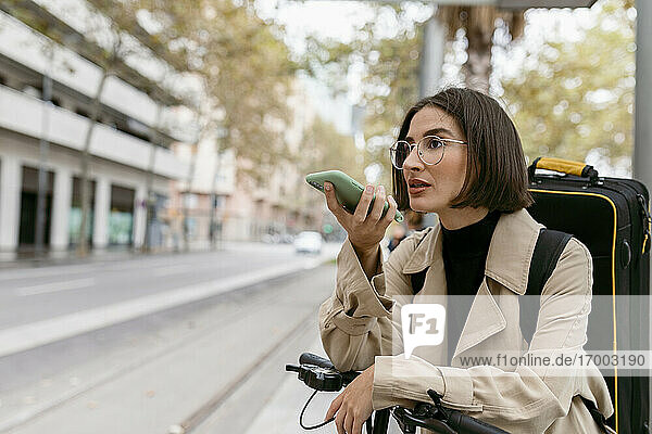 Schöne Frau mit Instrumentenkoffer und elektrischem Tretroller  die mit ihrem Handy telefoniert  während sie in der Stadt steht