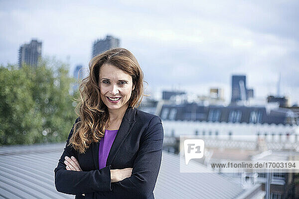 Selbstbewusst lächelnde Unternehmerin  die mit verschränkten Armen auf einer Terrasse vor dem Himmel steht