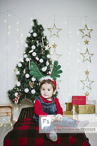 Niedlicher Junge mit gehörntem Stirnband  der an einem Weihnachtsbaum zu Hause sitzt