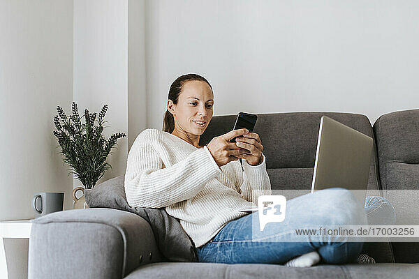 Frau schreibt eine SMS über ihr Smartphone  während sie mit ihrem Laptop auf dem Sofa sitzt