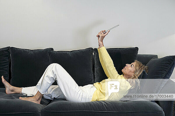 Reife Frau mit erhobenen Händen  die ein digitales Tablet benutzt  während sie zu Hause auf dem Sofa liegt