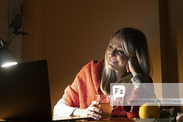 Smiling woman drinking tea while listening to video call on laptop at home