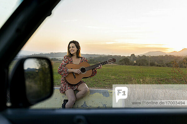 Lächelnde junge Frau spielt Gitarre aus dem Autofenster bei Sonnenuntergang gesehen