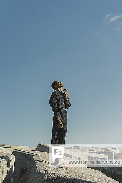 Junger Mann in schwarzem Kaftan steht auf Betonblöcken unter blauem Himmel