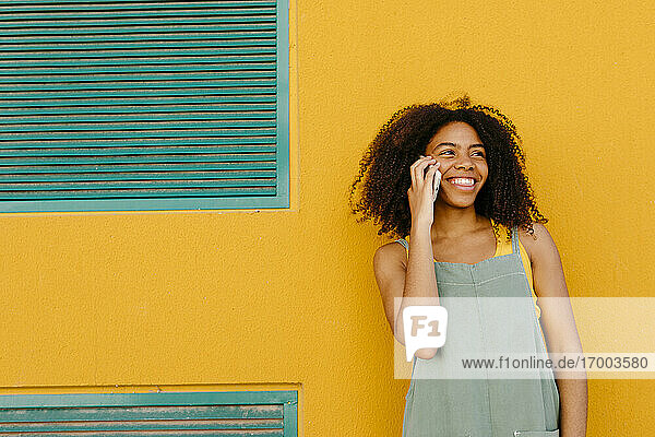 Porträt einer fröhlichen jungen Frau in Latzhose vor einer gelben Wand  die am Telefon spricht