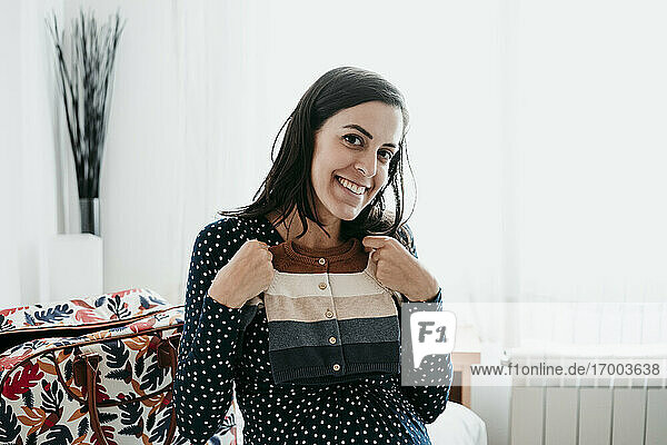 Happy pregnant woman holding baby clothes at home