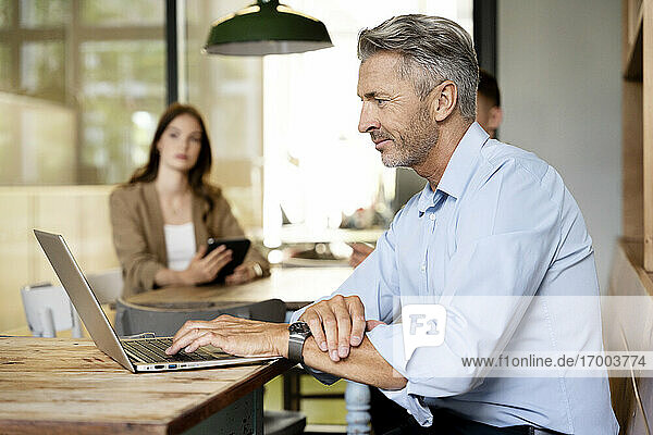 Älterer Geschäftsmann  der an einem Laptop arbeitet  während er mit Kollegen im Hintergrund im Büro sitzt