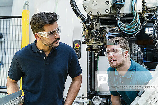 Nachdenkliche männliche Mitarbeiter mit Schutzbrillen  die Maschinen analysieren  während sie in der Industrie stehen