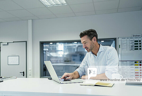 Männlicher Fachmann  der an einem Laptop arbeitet  während er im Sitzungssaal eines Fabrikbüros sitzt