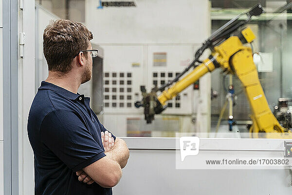 Männlicher Fabrikarbeiter mit verschränkten Armen  der einen Roboterarm durch ein Fenster betrachtet  während er in einer Fabrik steht
