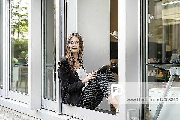 Junge Geschäftsfrau mit digitalem Tablet  die wegschaut  während sie an der Bürotür sitzt