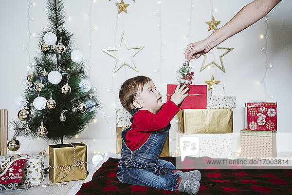 Mutter schenkt einem kleinen Jungen  der zu Hause auf einer Decke sitzt  eine Weihnachtskugel