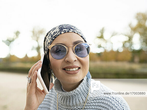 Junge Frau mit Kopftuch und Sonnenbrille lächelt  während sie im Freien mit einem Mobiltelefon spricht