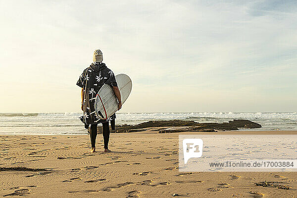 Mann hält Surfbrett auf Sand und schaut auf das Meer gegen den Himmel