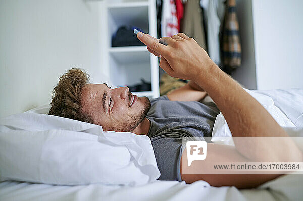 Junger Mann benutzt sein Smartphone  während er auf dem Bett im Schlafzimmer liegt