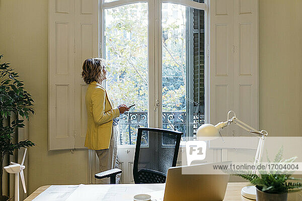 Unternehmerin schaut durch das Fenster und hält ihr Smartphone im Büro