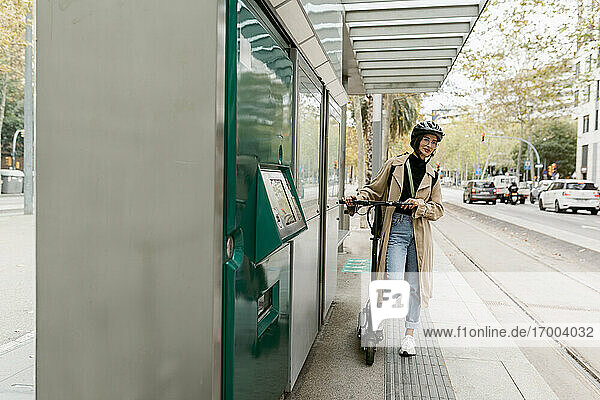 Frau mit Fahrradhelm  die mit einem Elektroroller an einer Straßenbahnhaltestelle in der Stadt wartet